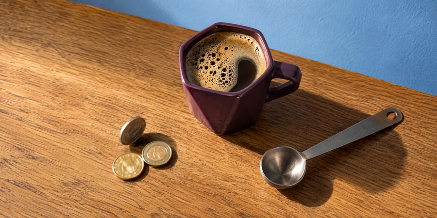 Taza de café con espuma junto a unas monedas de euro y una cuchara dosificadora sobre una mesa de madera